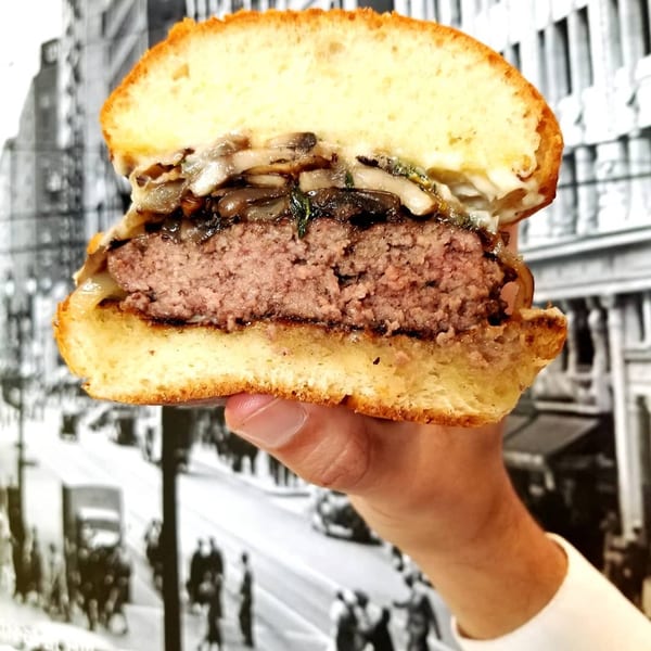 A hand holds a sliced burger with a thick patty, melted cheese, and sautéed mushrooms.