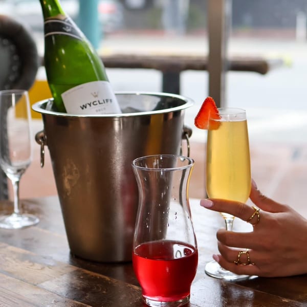 A Wycliff Brut champagne bottle in an ice bucket, with a mimosa and cranberry juice on a wooden table.