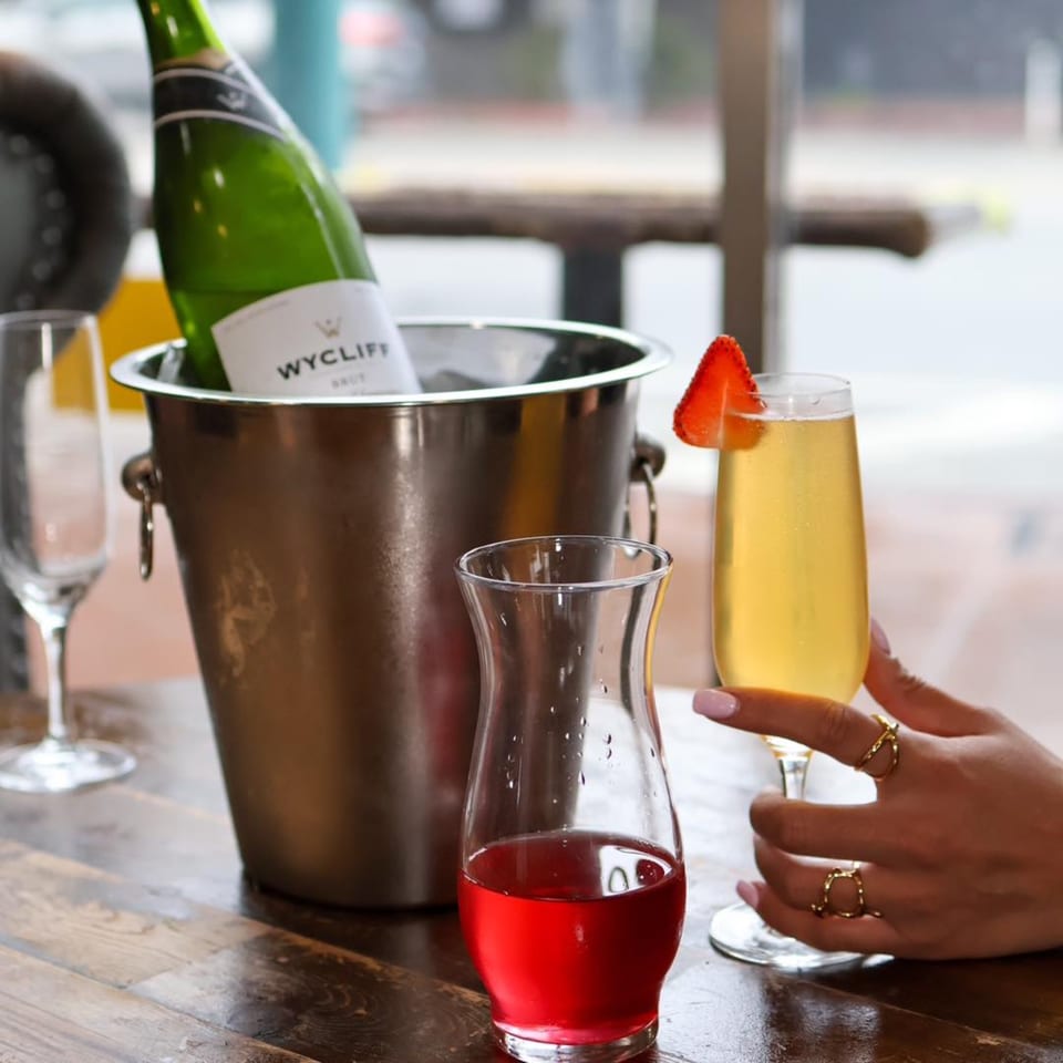 A Wycliff Brut champagne bottle in an ice bucket, with a mimosa and cranberry juice on a wooden table.