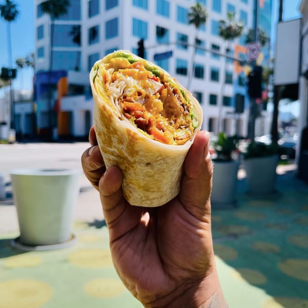 Hand holding a breakfast burrito filled with eggs, bacon, and cheese, with a city street in the background.